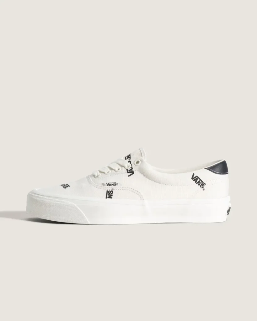 Side profile of the white Vans OTW low-top sneaker showcasing the all-over black Vans logo print, black heel tab, white canvas upper, and clean white rubber midsole