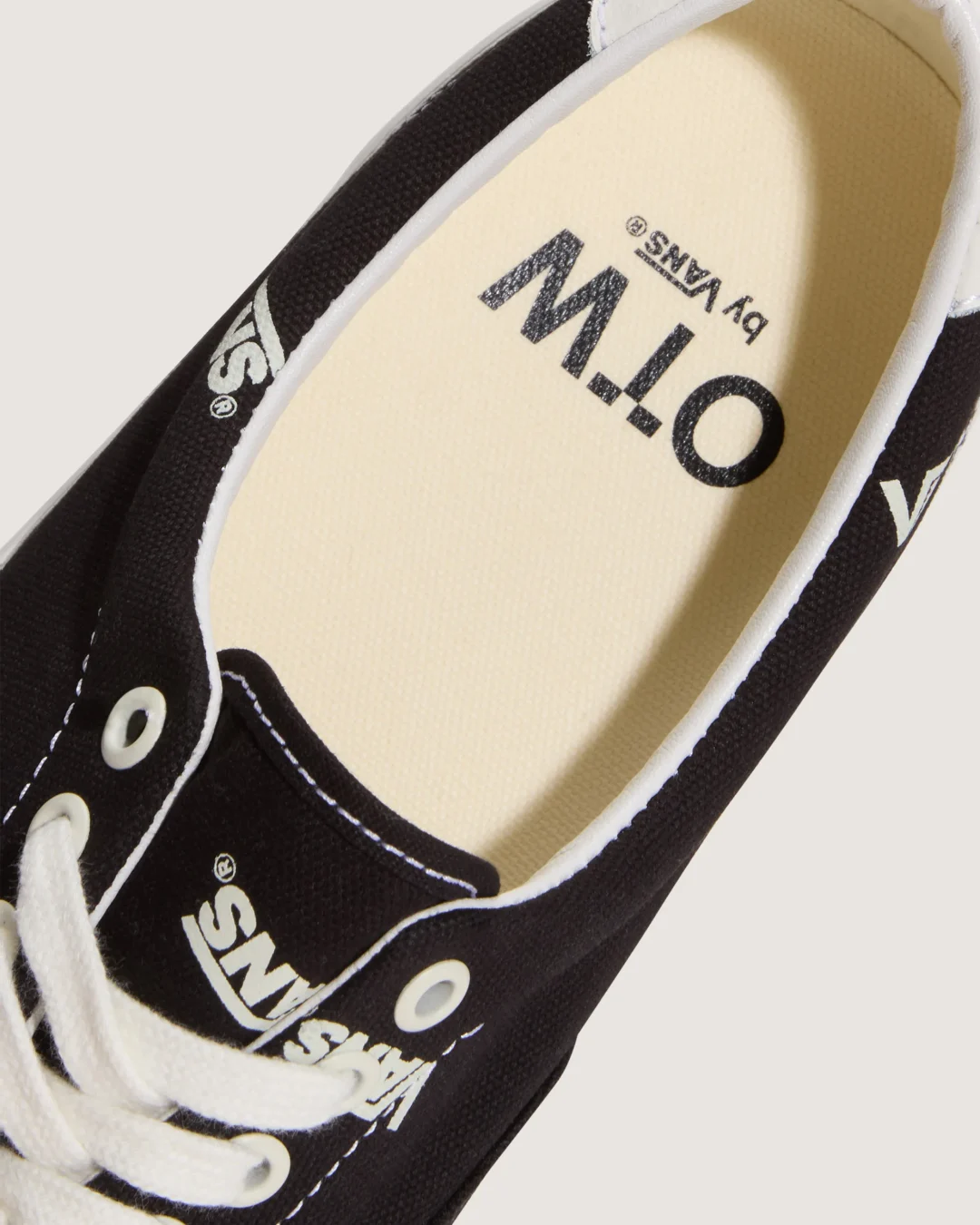 Close-up of the Vans OTW low-top sneaker showing the cream-colored cushioned insole with bold 'OTW by Vans' branding, surrounded by a black canvas upper featuring repeating Vans logo graphics, white eyelets, and white laces.