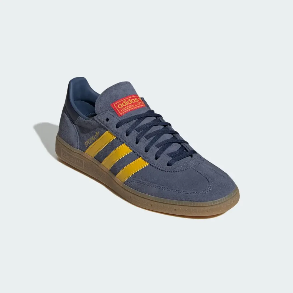 Gray-blue suede adidas Handball Spezial shoe shown from a front three-quarter angle, featuring bright yellow Three Stripes, navy laces, a red adidas tongue label, and a translucent gum rubber sole against a light gray background