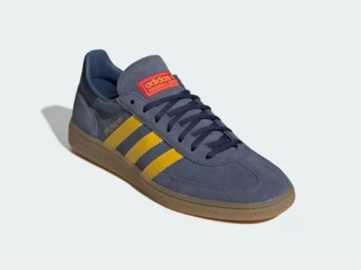 Gray-blue suede adidas Handball Spezial shoe shown from a front three-quarter angle, featuring bright yellow Three Stripes, navy laces, a red adidas tongue label, and a translucent gum rubber sole against a light gray background