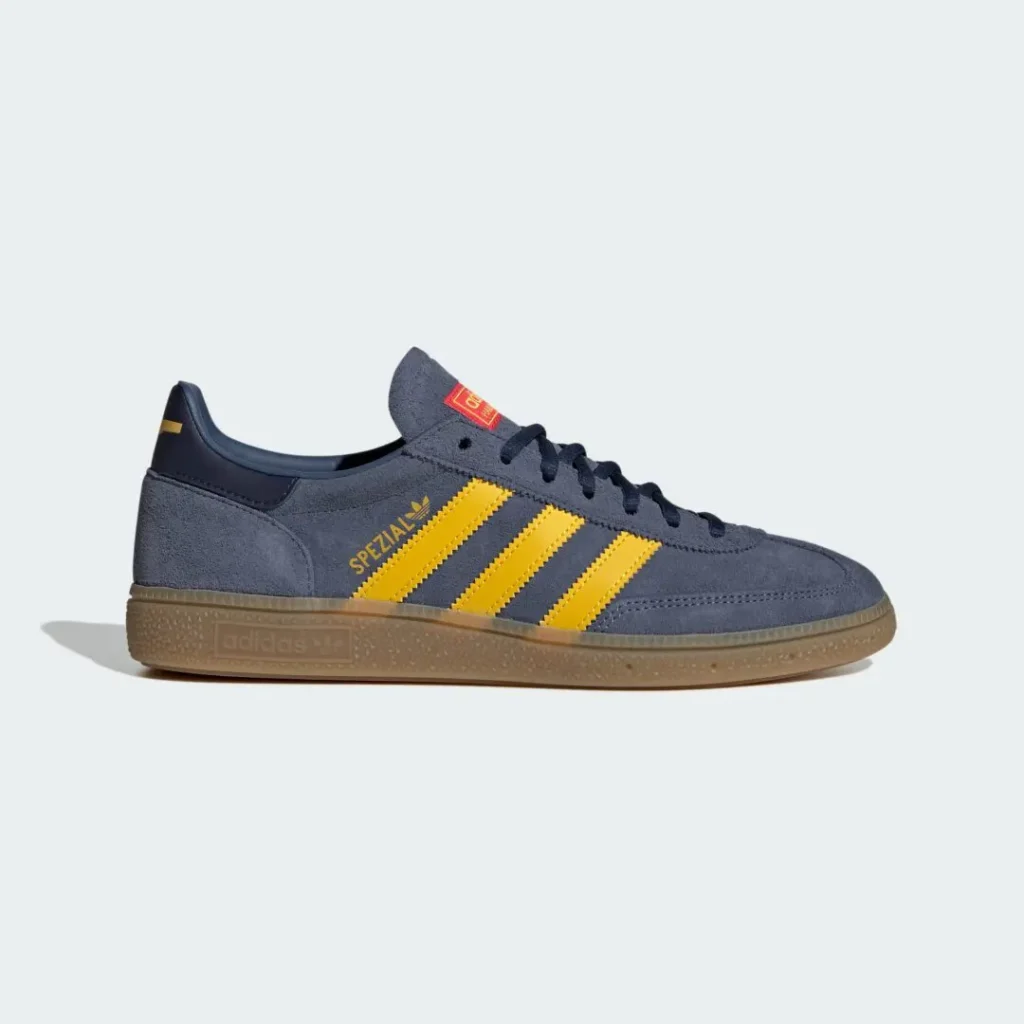 Lateral side profile of the adidas Handball Spezial in gray-blue suede with yellow Three Stripes, gold Spezial branding, navy heel tab, and a classic gum sole on a clean studio backdrop