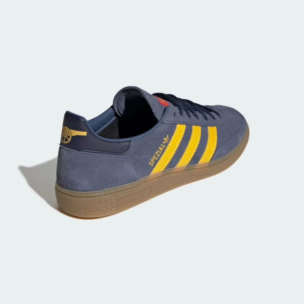 Rear three-quarter angle of the adidas Handball Spezial showcasing the suede heel, yellow Three Stripes, gold Spezial logo, navy leather heel tab with a yellow Arsenal cannon graphic, and gum outsole