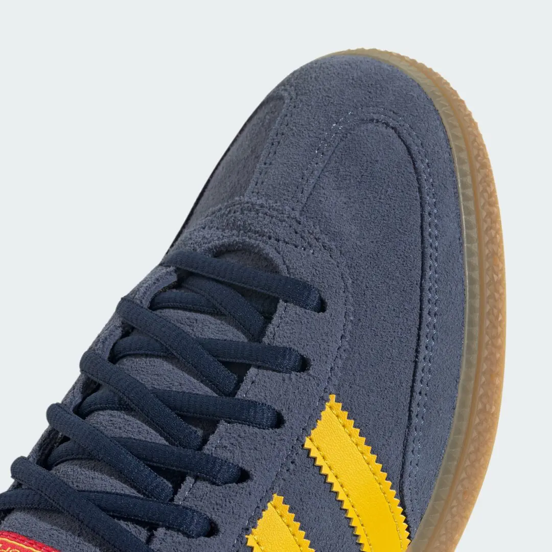 Close-up of the adidas Handball Spezial toe box highlighting the textured gray-blue suede upper, tonal stitching, navy flat laces, bright yellow leather Three Stripes, and translucent gum sole against a light gray studio background.