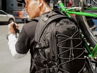 A person carries a green bicycle over one shoulder while wearing a black technical backpack with reflective bungee cord lacing, multiple compression straps, mesh side pockets, and subtle PORTER branding in an urban street set