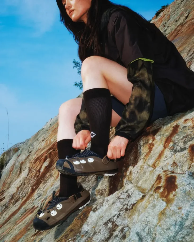 A woman sits on a rocky slope adjusting an olive Helinox trail clog while wearing black crew socks and lightweight technical outerwear. The image highlights the adjustable heel strap, quick-lace system, and outdoor-ready style