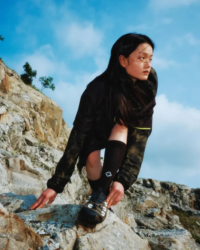 A woman climbs across a rocky outcrop wearing Helinox trail clogs, black performance socks, and lightweight hiking apparel. The low-angle composition emphasizes grip, flexibility, and movement across uneven terrai