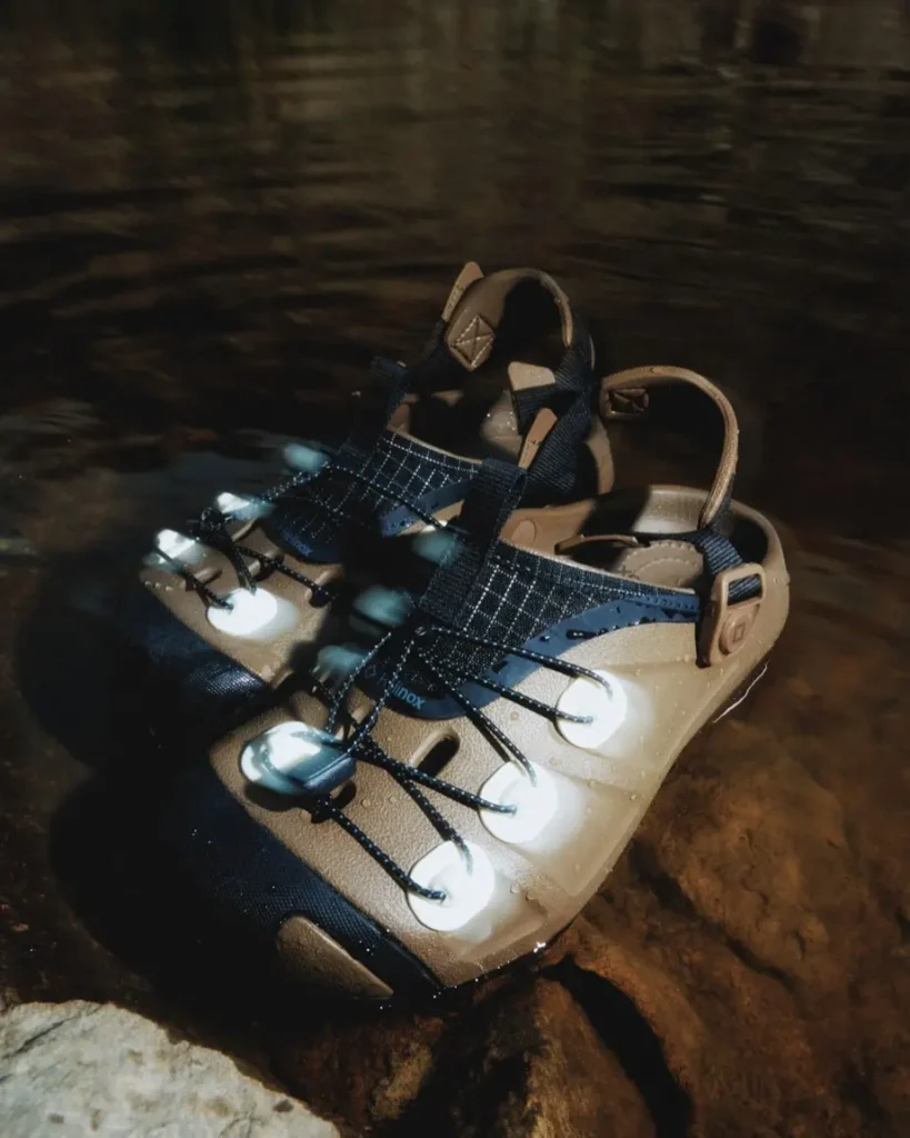 Pair of olive Helinox trail clogs resting on wet rocks beside shallow water. Water droplets cover the molded upper while the reflective white lace anchors and technical lacing system stand out in the close-up product shot