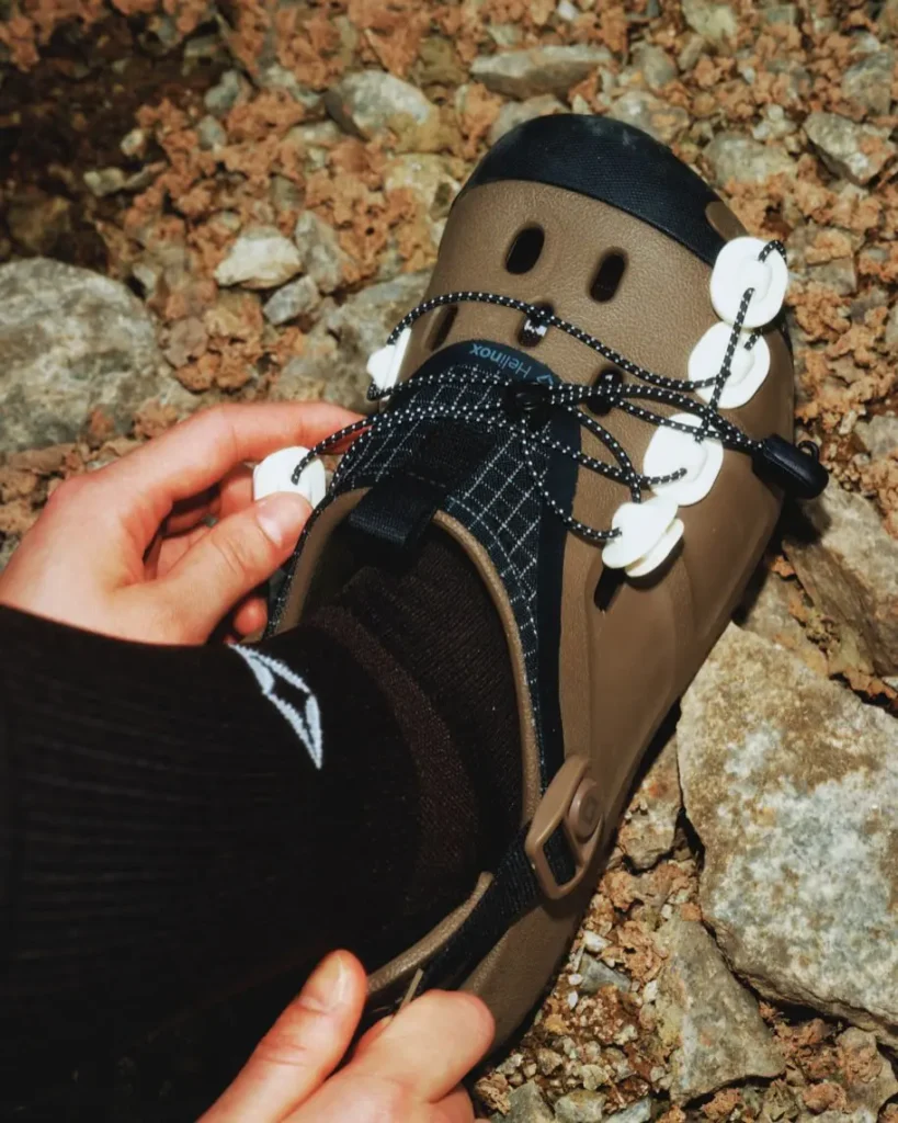 Close-up of an olive Helinox trail clog being adjusted by hand on rocky ground. The image showcases the molded shell, reflective lace anchors, quick-lace system, adjustable heel strap, and protective toe cap