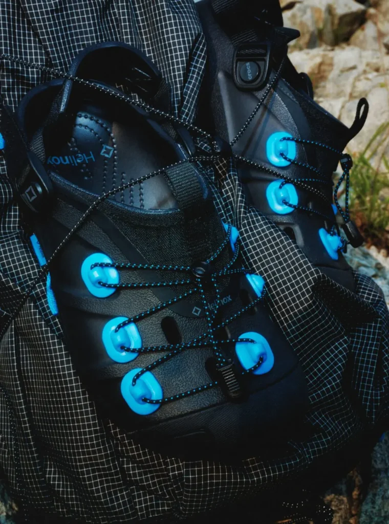 Detailed close-up of a pair of black Helinox trail clogs secured to a technical backpack. Bright blue lace anchors, reflective cord lacing, molded construction, and Helinox branding are highlighted against ripstop fabric