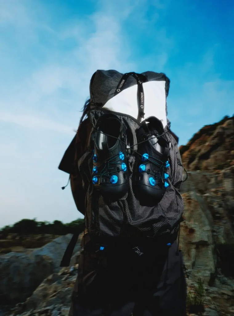 Technical hiking backpack displayed outdoors with a pair of black Helinox trail clogs attached to the front. The composition highlights lightweight adventure gear against a rocky landscape and blue evening sky