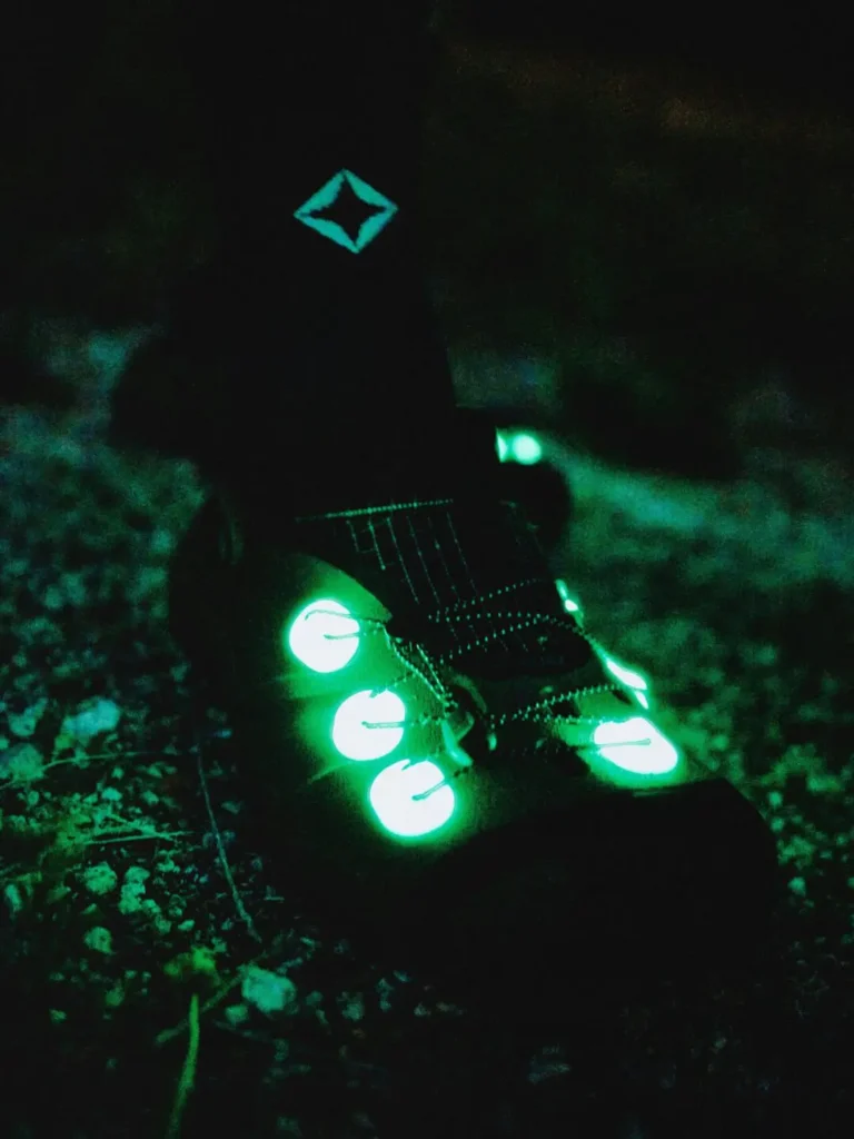Two trail runners move across a grassy landscape at night, photographed with bright blue light flares that create a dramatic long-exposure effect. Both wear Helinox outdoor apparel and technical footwear designed for after-dark adventure