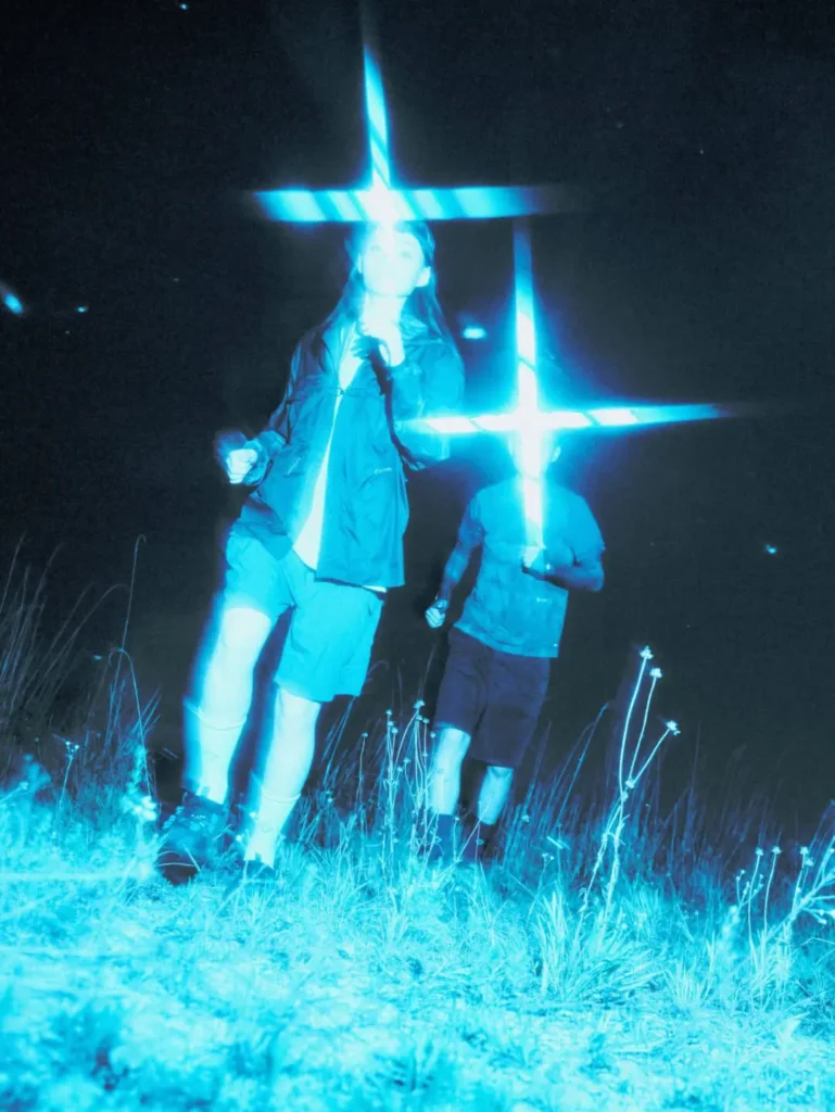 Rear view of three people standing on a gravel trail wearing Helinox socks and rugged trail clogs. The glow-in-the-dark lace anchors and heel details emit a soft green glow against the dim evening landscape