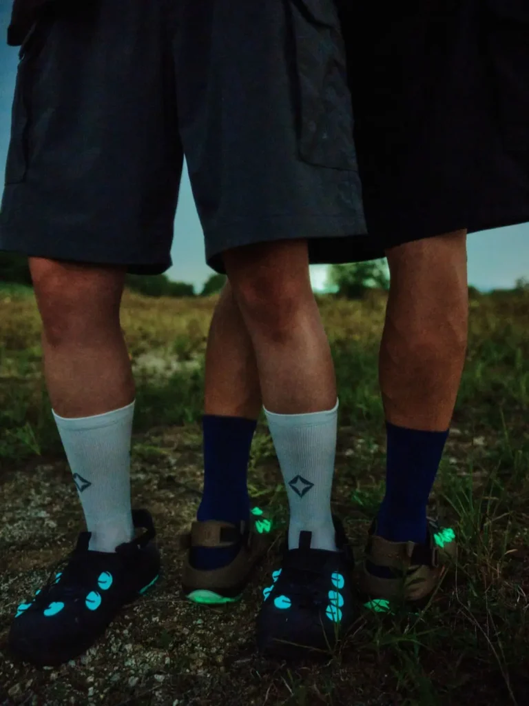 Close-up of a person standing on a rock at dusk wearing black Helinox crew socks and olive trail clogs with reflective white lace anchors, black toe protection, and a technical lace system designed for outdoor terrain
