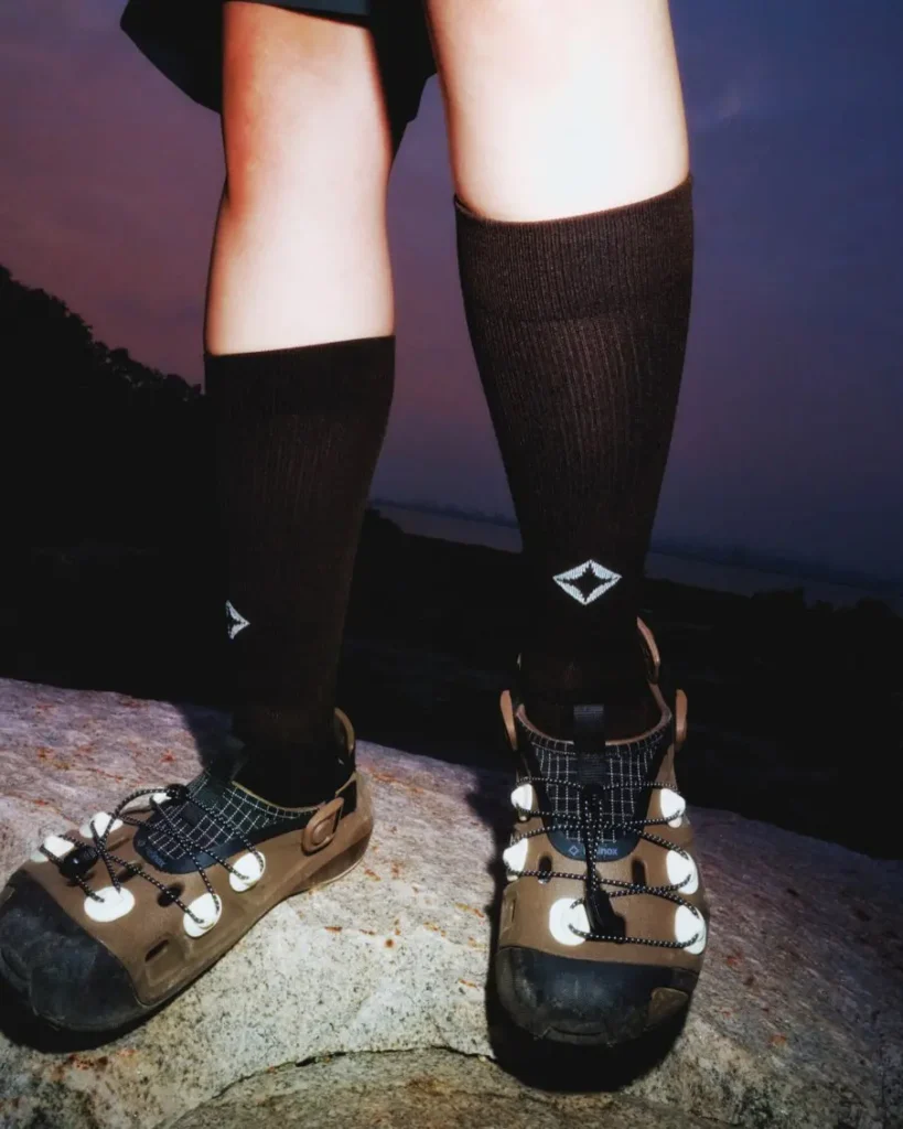 Nighttime action photograph of a hiker illuminated by a bright headlamp while walking through rocky terrain. The glowing Helinox trail clogs and blue-tinted lighting emphasize visibility and show in low-light condition