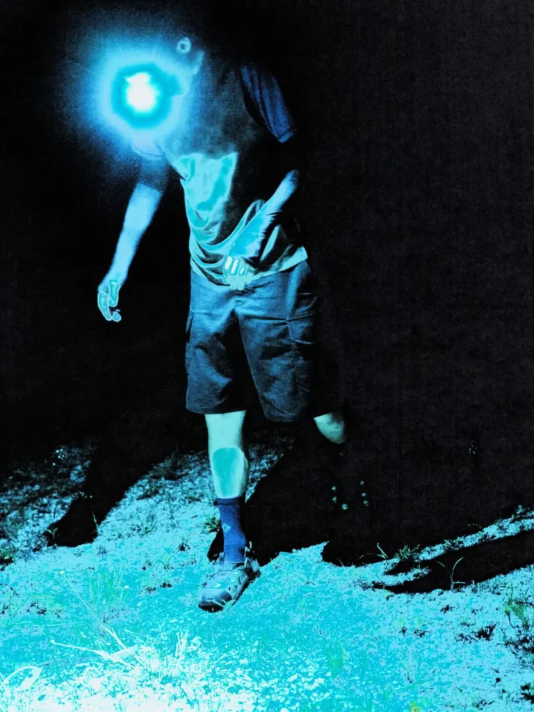 Dramatic close-up of a Helinox trail clog beside a bright campfire. The glow-in-the-dark lace anchors shine blue-white while the flames cast warm light across the rugged outdoor footwear and surrounding grave