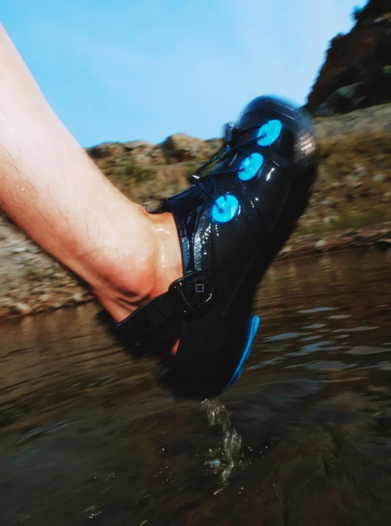 Close-up of a black Helinox trail clog splashing through shallow water. Bright blue glow-in-the-dark lace anchors contrast against the wet upper and rocky stream, highlighting the shoe's outdoor-ready construction