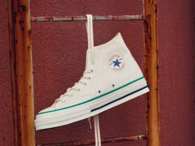 Cream Converse Chuck Taylor All Star high-top shoe suspended from a weathered metal ladder, featuring a subtle tonal graphic canvas upper, green foxing stripe, blue heel stripe, and classic ankle patch