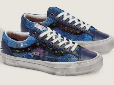 Pair of Vans Old Skool low-top shoe displayed at an angle, featuring blue Harris Tweed wool uppers, navy leather overlays, white laces, distressed foxing, jewel embellishments along the side stripe, silver cone studs at the heel, and embroidered pink detail