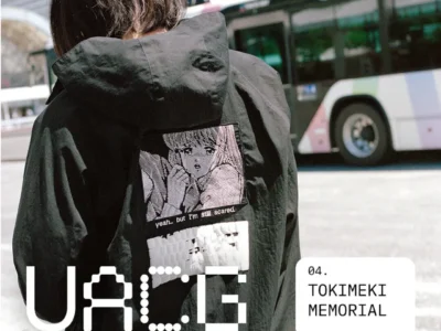 Person wearing a black hooded jacket with a large manga-inspired back graphic and subtitle text, photographed on a city street with a bus in the background