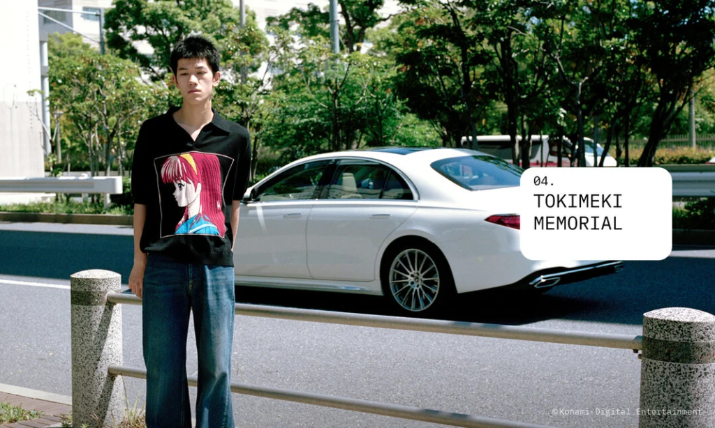 Model standing beside a city street wearing a black knit polo featuring a large embroidered anime character portrait on the front with relaxed blue jean