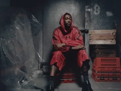 Campaign portrait of a model seated in a dim industrial room wearing a red adidas Originals by Taqwa Bint Ali hoodie and matching sculpted shorts with black shoe