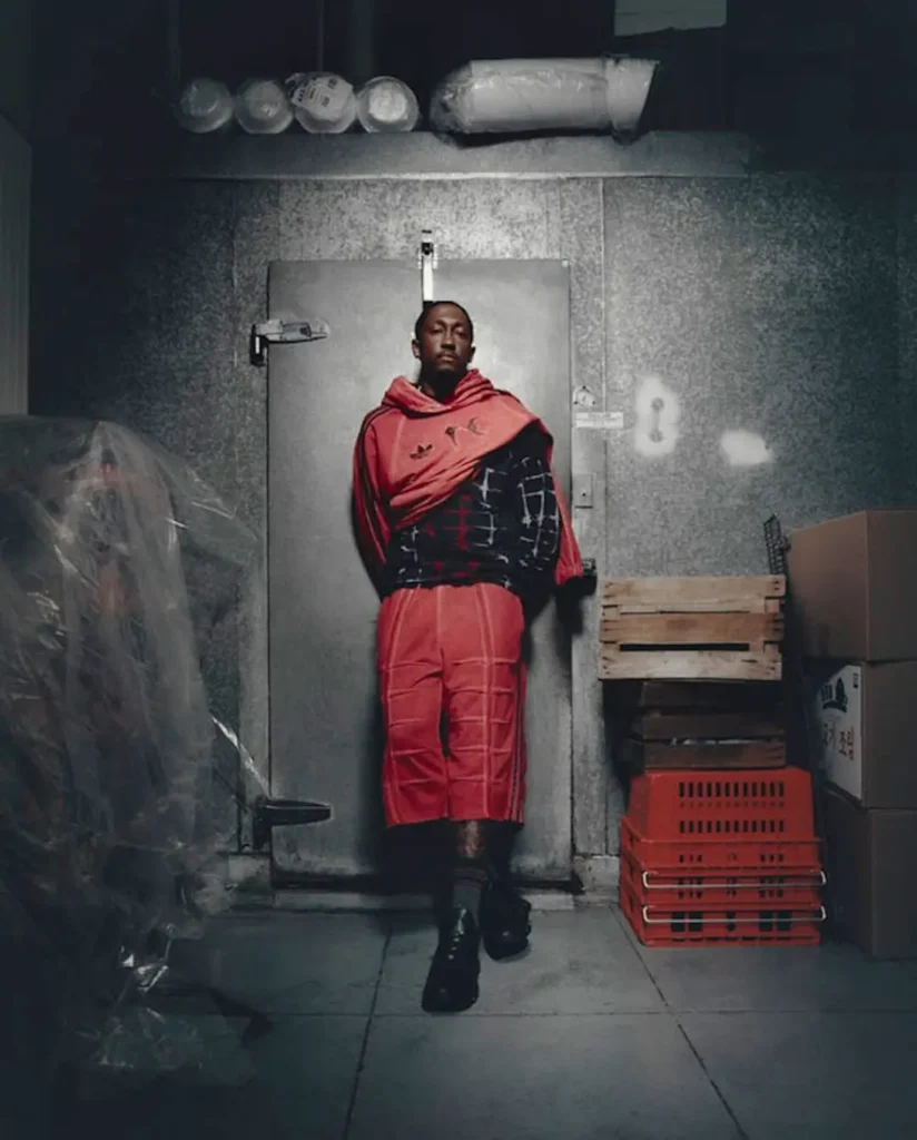 Campaign portrait of a model standing in an industrial setting wearing a red adidas Originals by Taqwa Bint Ali hoodie and matching sculpted shorts with black footwear