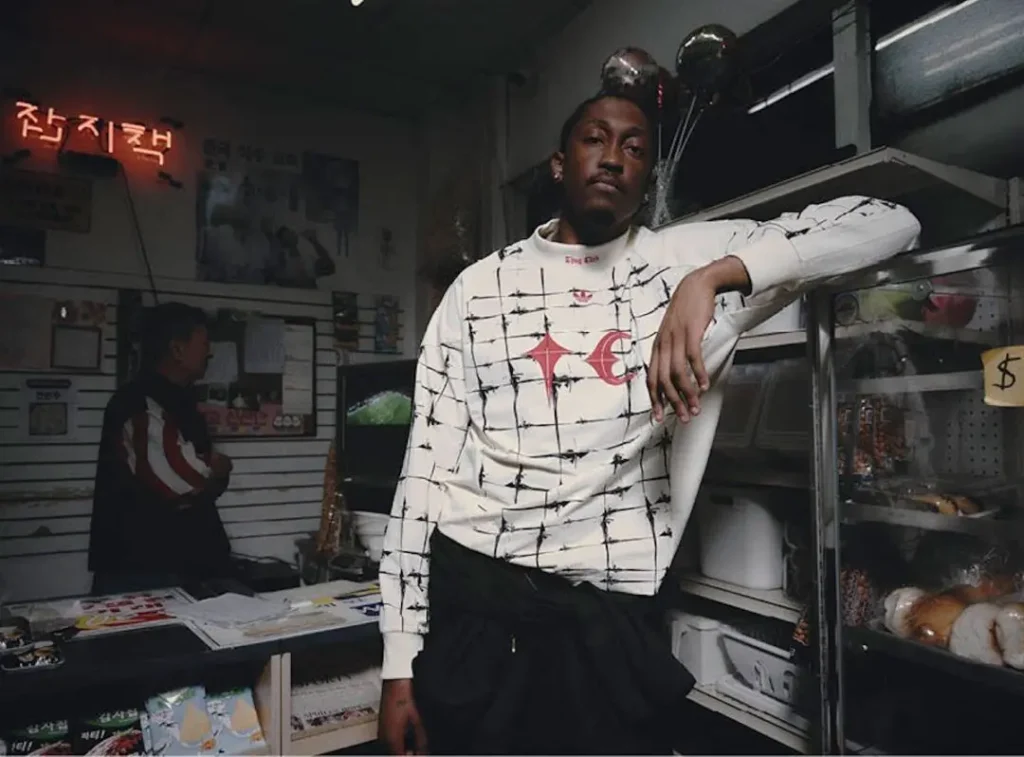 Campaign portrait of a model inside a neighborhood convenience store wearing a white adidas Originals by Taqwa Bint Ali long-sleeve top with black grid graphics and co-branded chest logo