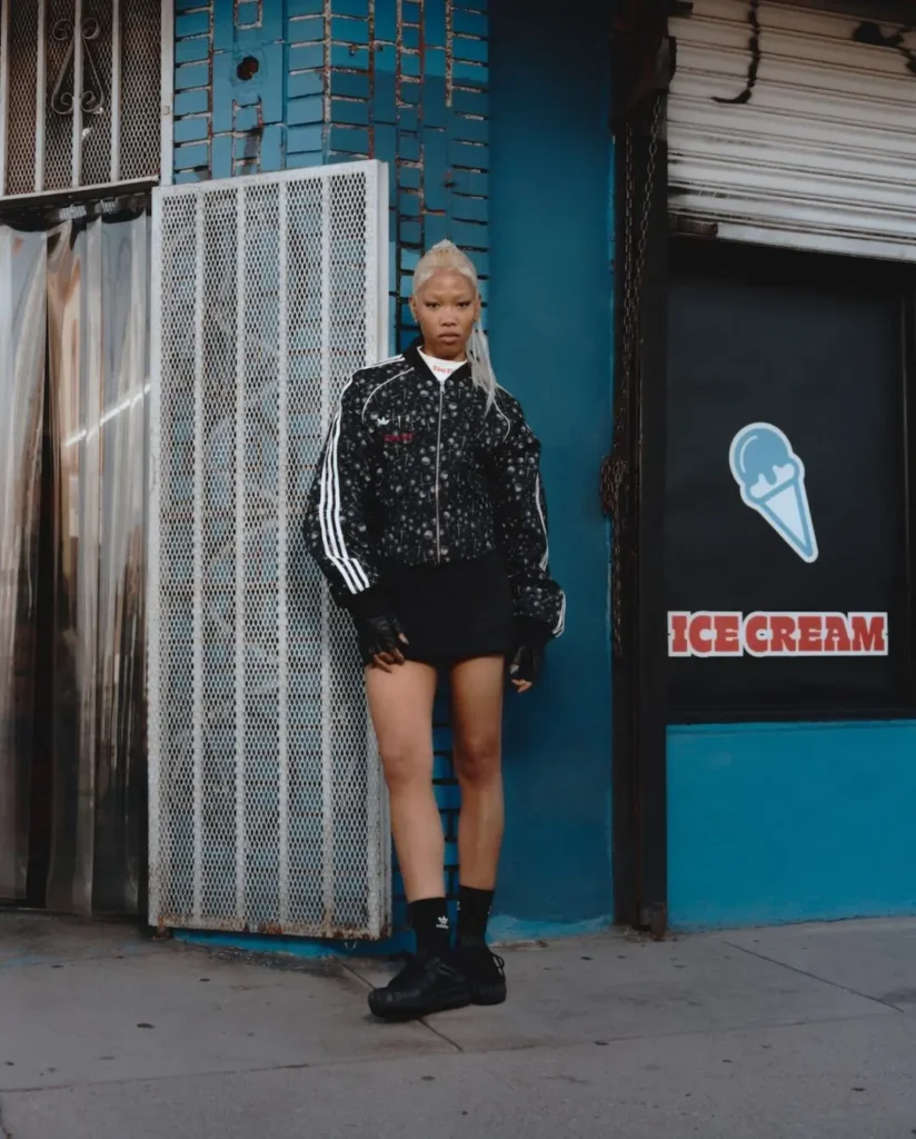 Campaign portrait of a model standing outside a storefront wearing a black adidas Originals by Taqwa Bint Ali track jacket with graphic print, black shorts, and black footwear