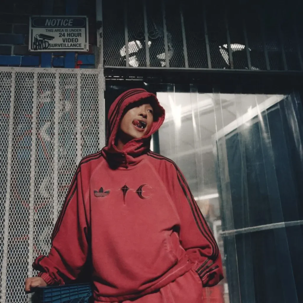 Campaign portrait of a model wearing a red adidas Originals by Taqwa Bint Ali hoodie with co-branded chest graphics outside an urban storefront