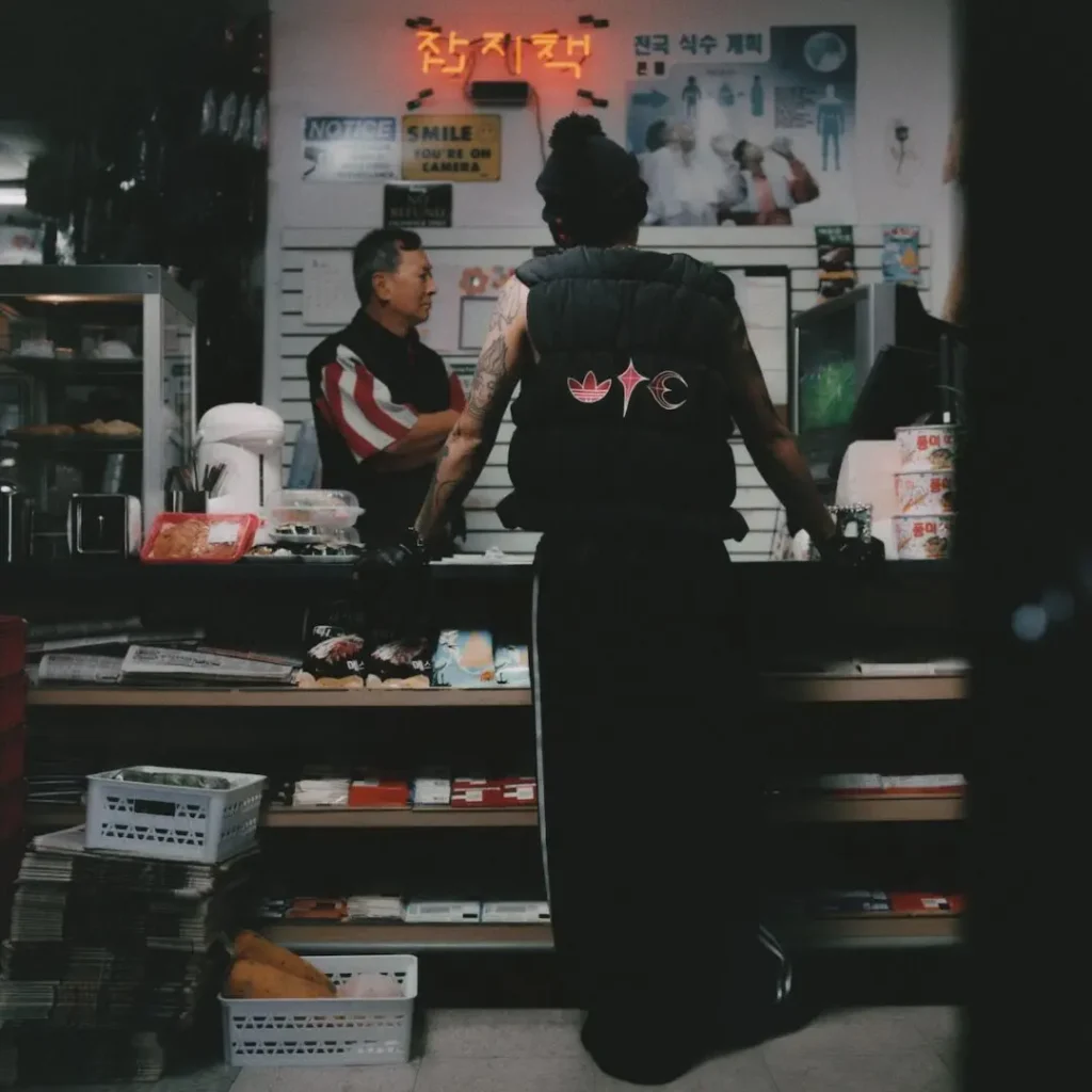 Campaign image of a model at a convenience store counter wearing a black adidas Originals by Taqwa Bint Ali padded vest with co-branded back graphic