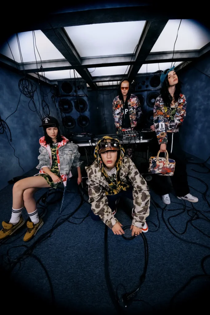 Four models posing in mixed camouflage jackets, denim, printed handbags, and streetwear around DJ equipment with hanging cables and oversized speaker