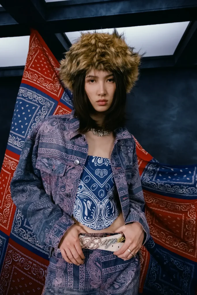 Model wearing a purple paisley-print denim jacket and matching pants with faux fur hat, blue patterned top, and decorative belt
