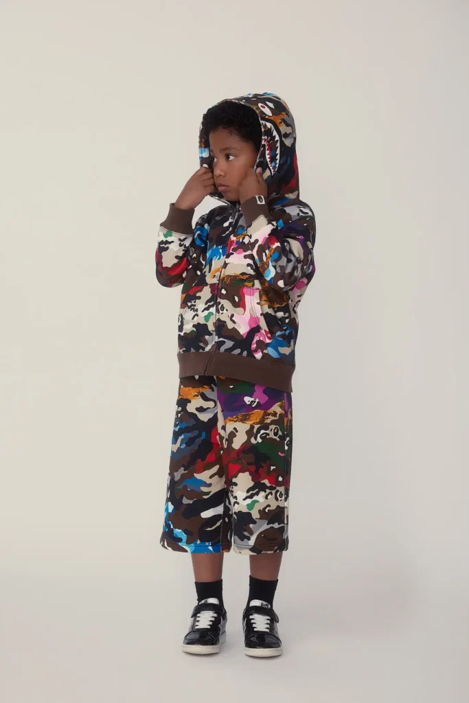 Child wearing a multicolor camouflage hoodie and matching shorts with black sneakers in a minimalist studio portrait