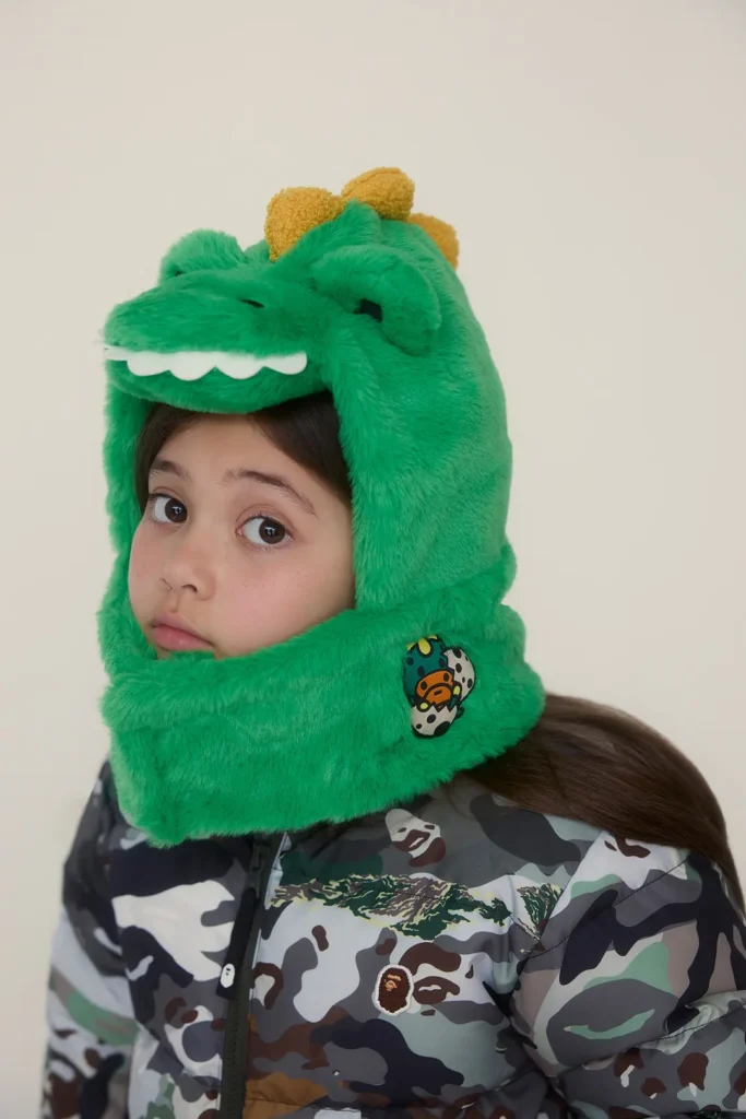 Child wearing a green dinosaur faux-fur hat and camouflage puffer jacket in a playful close-up portrait