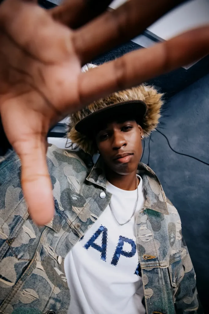 Model reaching toward the camera wearing a distressed camouflage denim jacket, white graphic T-shirt, furry trapper hat, and silver chain necklace