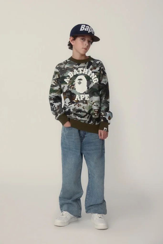 Child model wear a navy BAPE baseball cap, camouflage A Bathing Ape logo sweatshirt, relaxed blue jeans, and white sneakers in a minimalist studio portrait