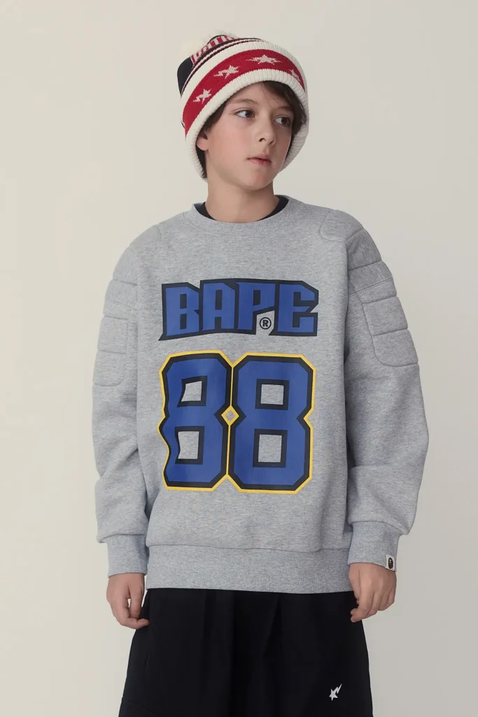 Child wearing a gray BAPE "88" sweatshirt with quilted sleeve panels, black shorts, and a red-and-white knit beanie in a casual studio portrait
