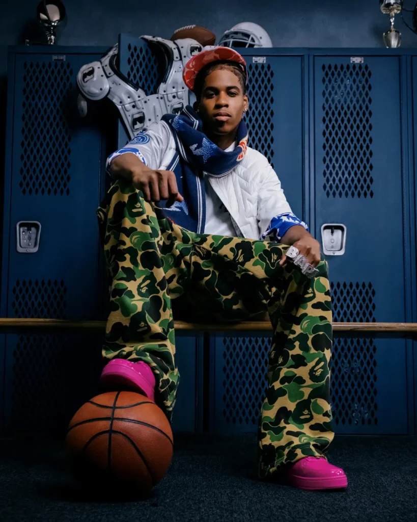 Model posing in a locker room wearing green camouflage BAPE pants, white quilted jacket, blue scarf, pink clogs, and orange camouflage cap with a basketball