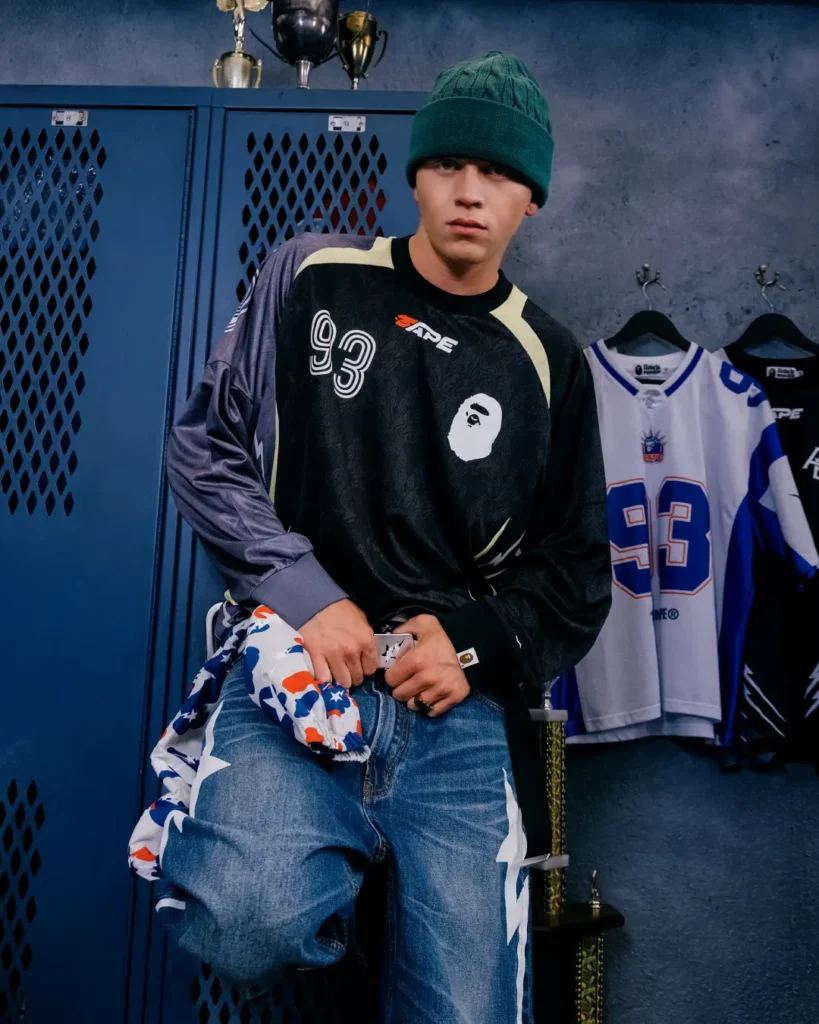 Model wearing a black BAPE football jersey with number 93, blue jeans, green knit beanie, and camouflage accessories in front of sports locker