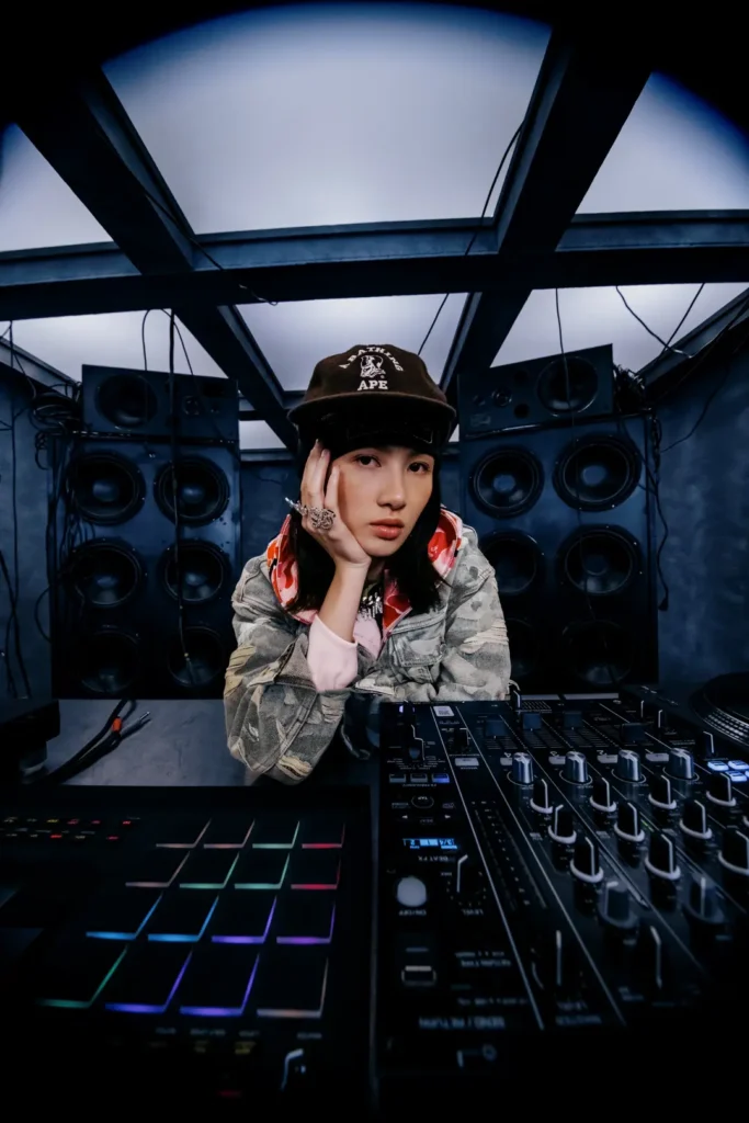 Model sitting behind DJ equipment wearing a camouflage denim jacket, black BAPE cap, pink hoodie, and silver rings in a music studio with large speaker
