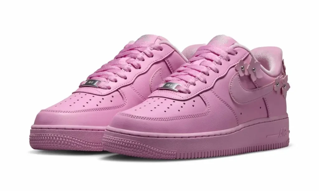 Three-quarter view of pastel pink Nike Air Force 1 sneakers featuring removable flower lace charms with silver stud center