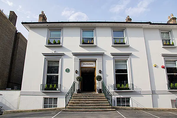 Exterior view of Abbey Road Studios, a white Georgian-style building with a central staircase leading to the main entrance. Symmetrical sash windows, black railings, and potted topiary frame the historic London recording studio beneath a bright blue sky.