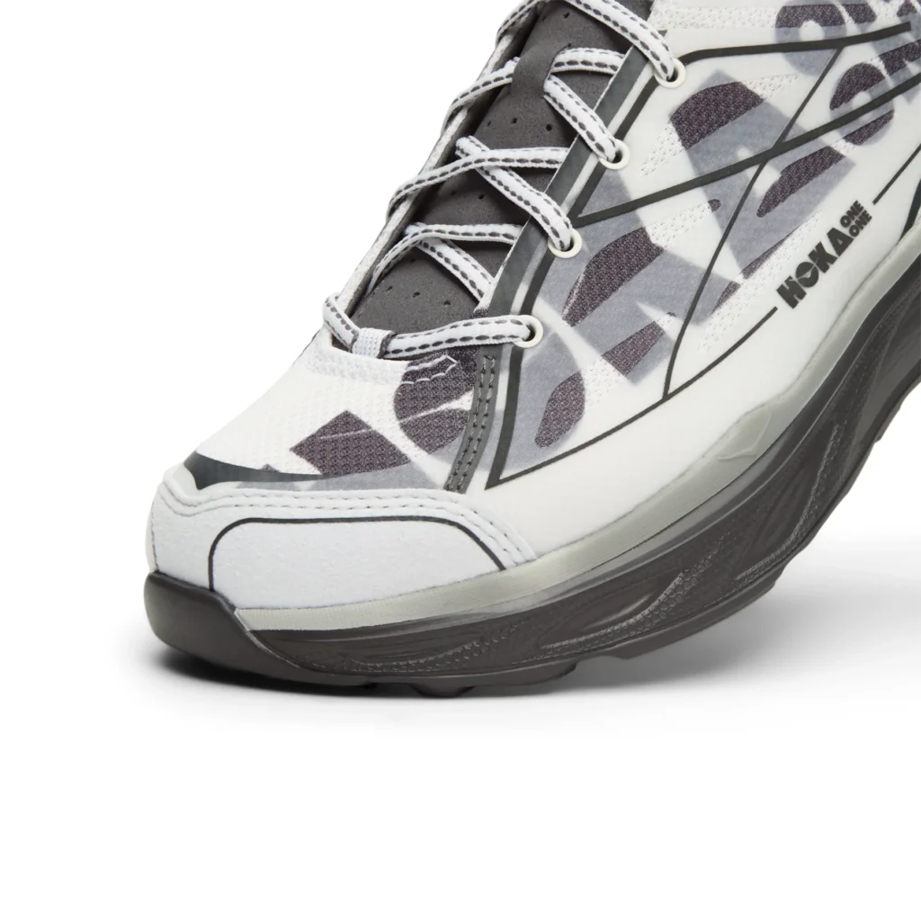 Close-up of the forefoot and lateral side highlighting the translucent layered upper, oversized printed HOKA letter, gray structural overlays, white stitched laces, reinforced toe bumper, and sculpted foam mid