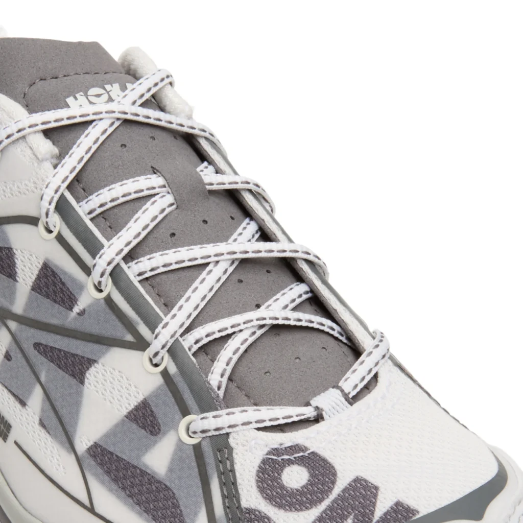 Close-up of the lacing system and tongue showing white contrast-stitched laces threaded through reinforced eyelets, a gray perforated tongue with HOKA branding, and translucent overlays framing the oversized side graphic