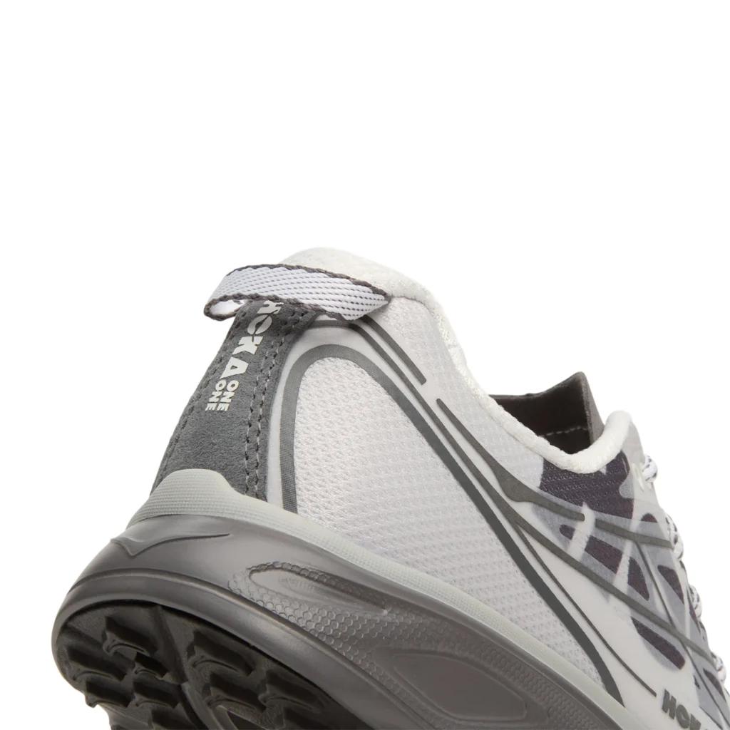 Close-up of the heel showcasing the textured white mesh upper, gray heel counter with vertical HOKA logo branding, pull tab, layered synthetic support lines, and the thick gray cushioned midsole above the black rubber outsole
