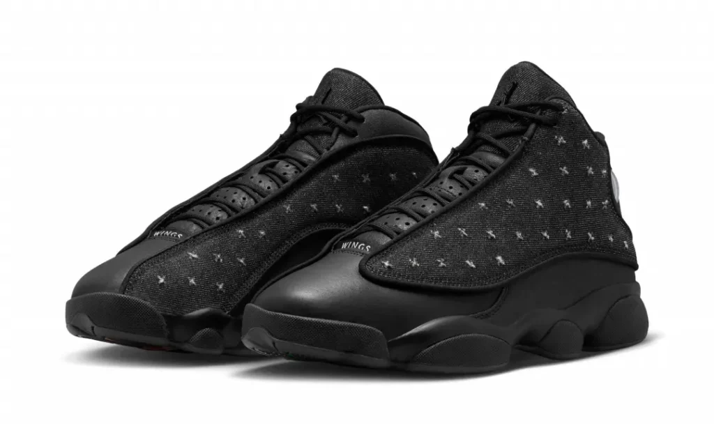 Air Jordan 13 "Wings" shoe in black feature distressed denim overlays, premium leather mudguards, and signature holographic ankle badges shown from a front three-quarter angle