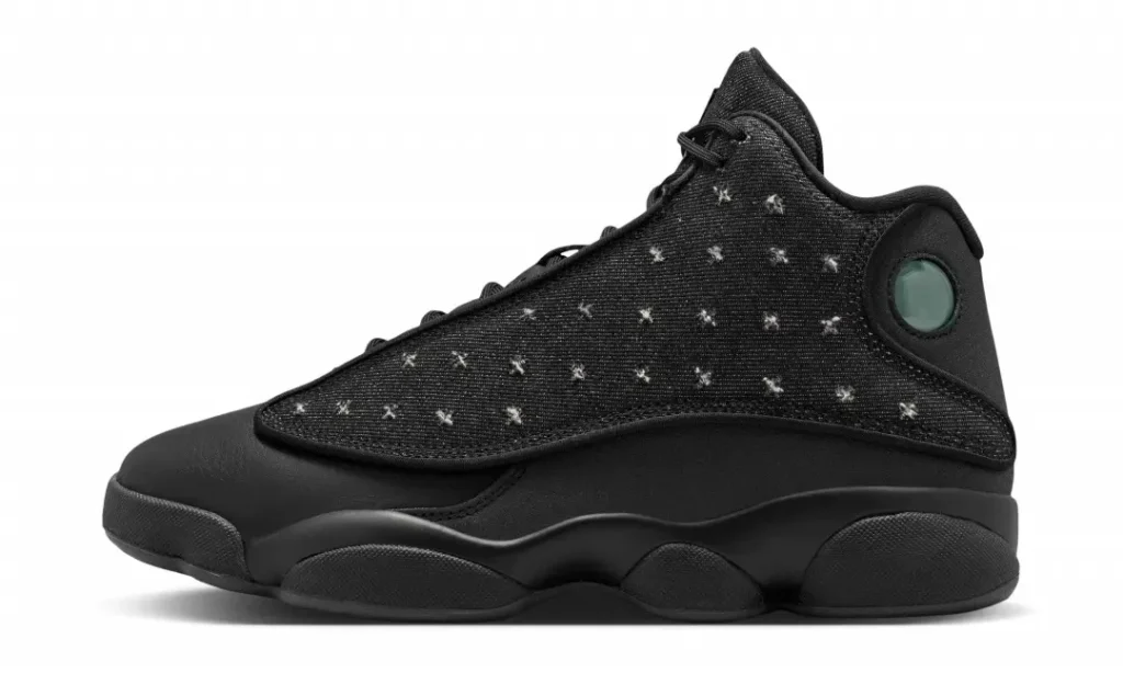 Lateral profile of the Air Jordan 13 "Wings" showcases black leather construction, distressed denim upper with faded cross-stitch detailing, and the iconic green hologram at the heel