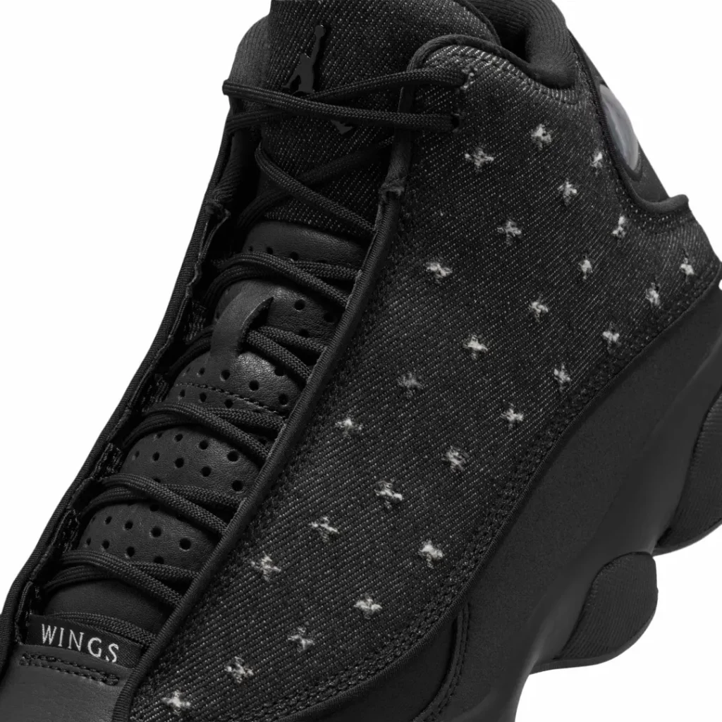 Lateral profile of the Air Jordan 13 "Wings" showcases black leather construction, distressed denim upper with faded cross-stitch detailing, and the iconic green hologram at the heel