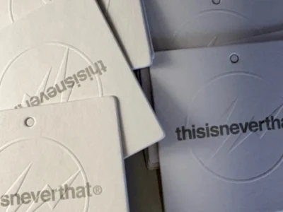 Stack of thisisneverthat branded hang tags in off-white and gray, featuring embossed circular graphics, lightning-inspired logo details, and minimalist typography arranged in an overlapping display