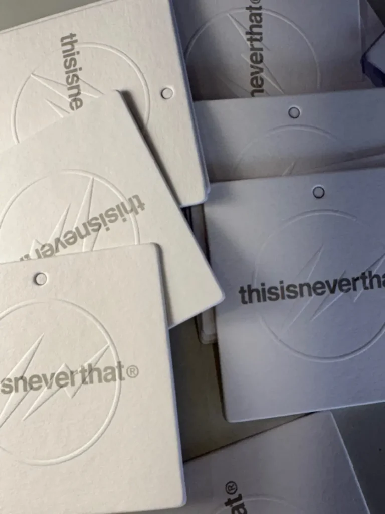 Stack of thisisneverthat branded hang tags in off-white and gray, featuring embossed circular graphics, lightning-inspired logo details, and minimalist typography arranged in an overlapping display